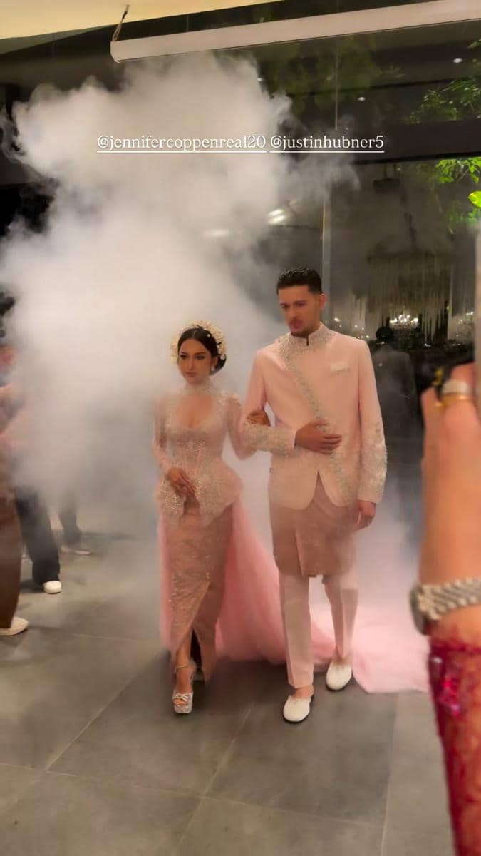 Pasangan pengantin mengenakan busana berwarna pink berjalan beriringan di acara wedding dinner dengan latar asap dekoratif.