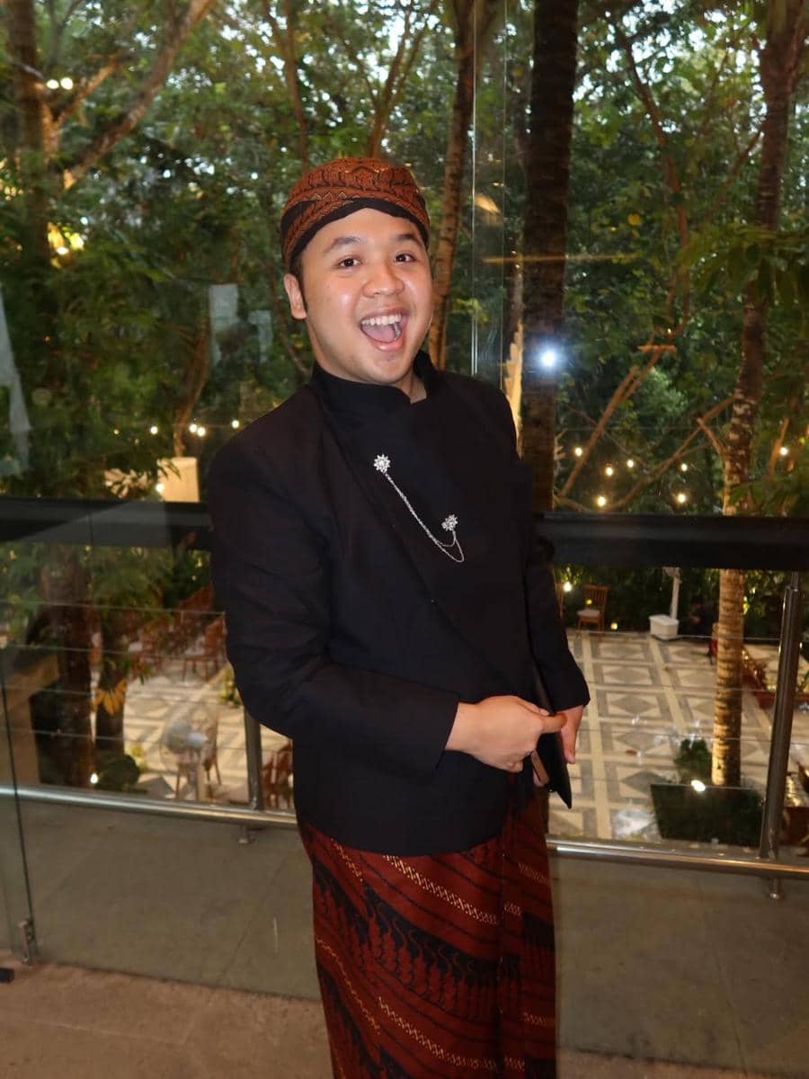 Seorang tamu pria mengenakan beskap hitam dan kain batik cokelat dengan blangkon, tersenyum lebar di acara akad pernikahan.