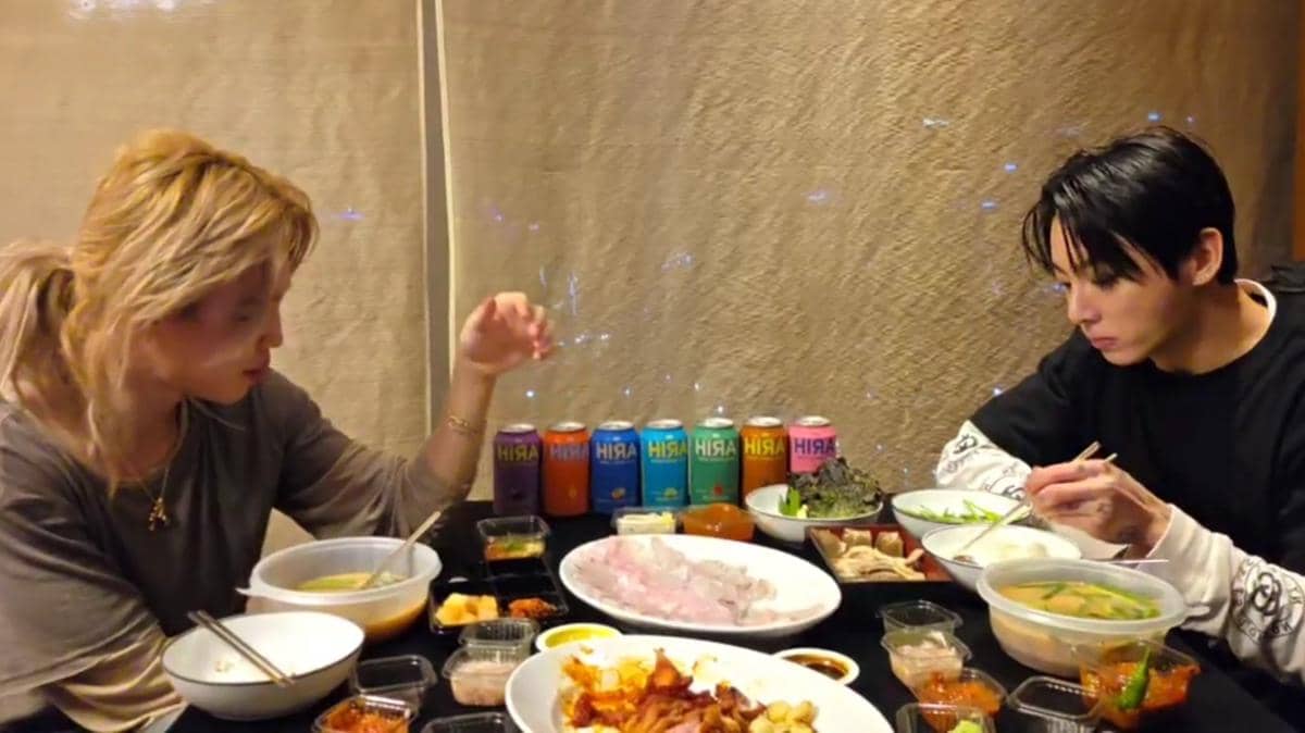 Dua orang duduk di meja makan menikmati hidangan Korea dengan berbagai lauk, minuman kaleng warna-warni, dan mangkuk sup di depan mereka.