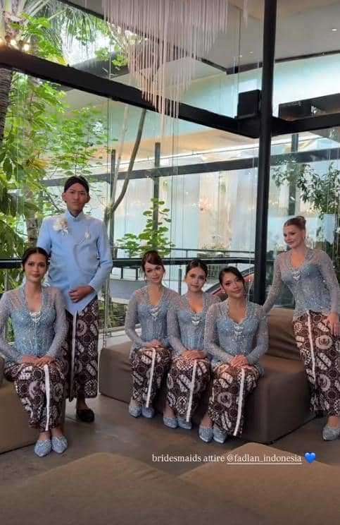 Enam orang bridesmaid mengenakan kebaya biru muda dan kain batik cokelat duduk dan berdiri di ruangan modern dengan pencahayaan alami.