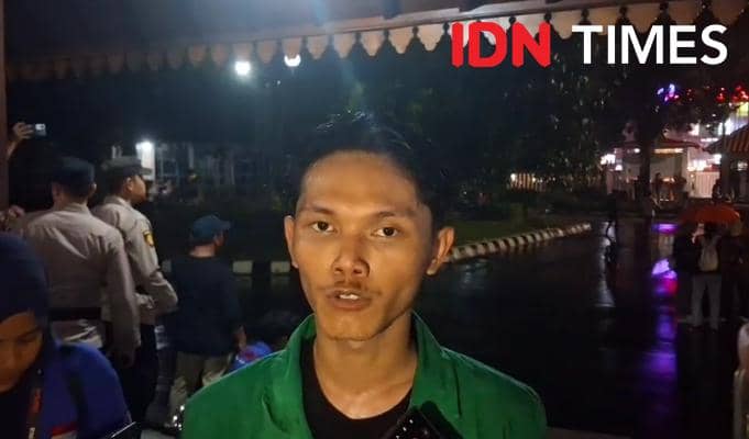 Seorang mahasiswa mengenakan jaket almamater hijau menyampaikan pernyataan di malam hari saat aksi evaluasi program pemerintah pusat.