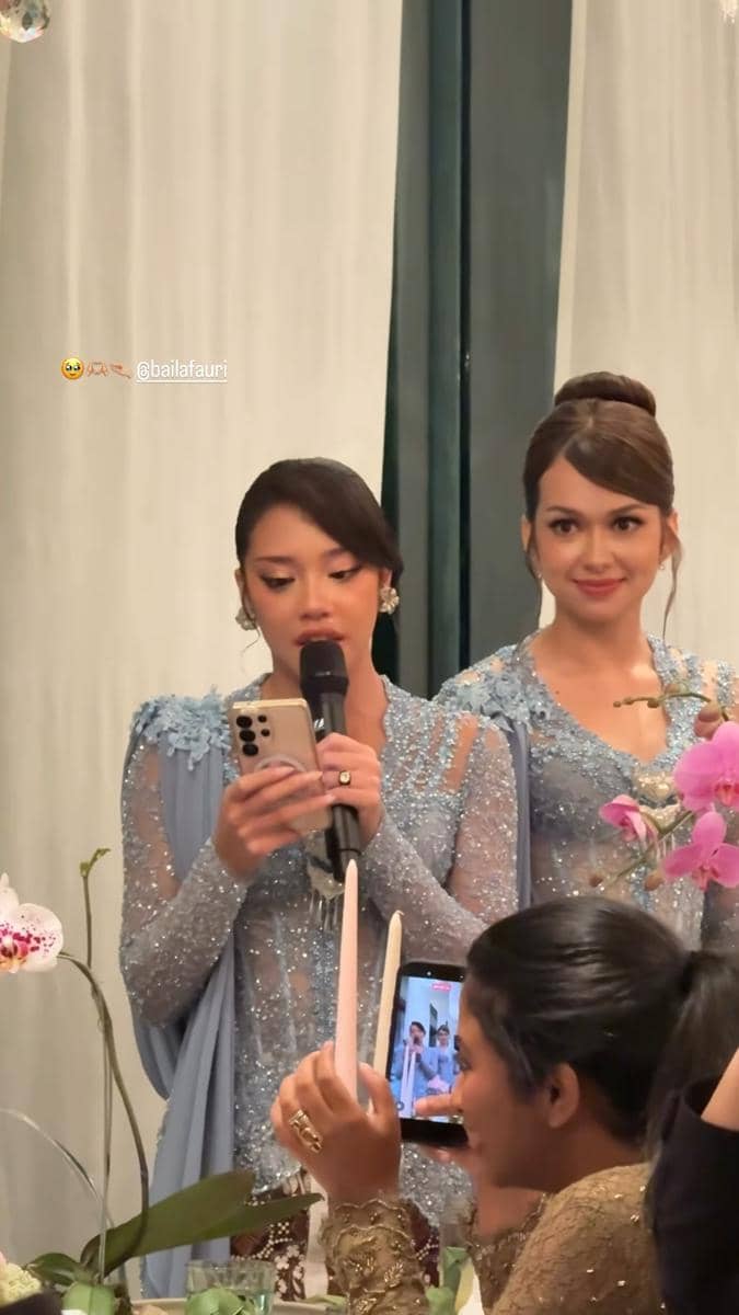 Dua wanita mengenakan gaun biru berhias payet sedang berdiri di acara makan malam pernikahan, salah satunya memegang mikrofon dan ponsel.