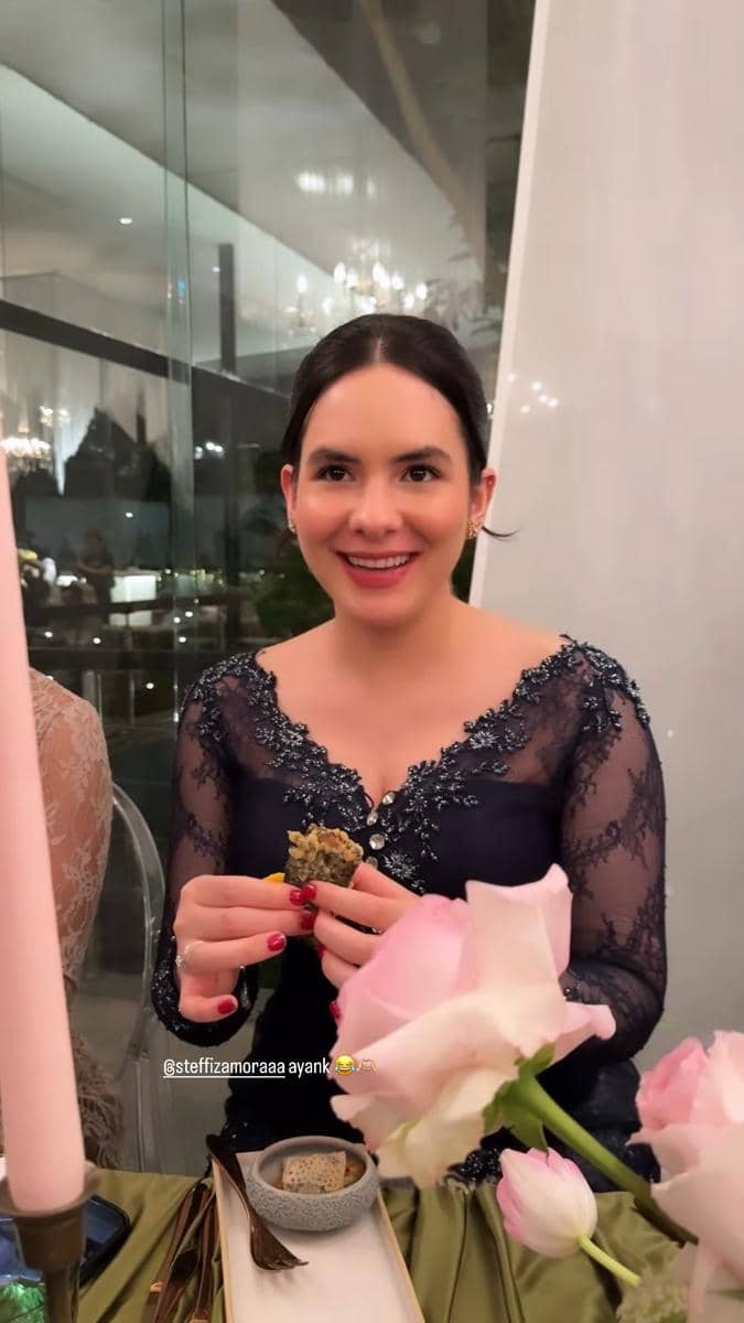 Seorang tamu mengenakan gaun biru navy berhias renda duduk di meja makan dengan dekorasi bunga saat acara akad Jennifer Coppen dan Justin Hubner.