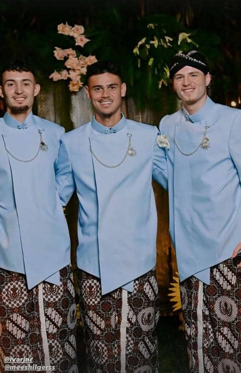 Tiga pria mengenakan busana adat Jawa berwarna biru muda dengan kain batik, tersenyum bersama di acara pernikahan malam hari.