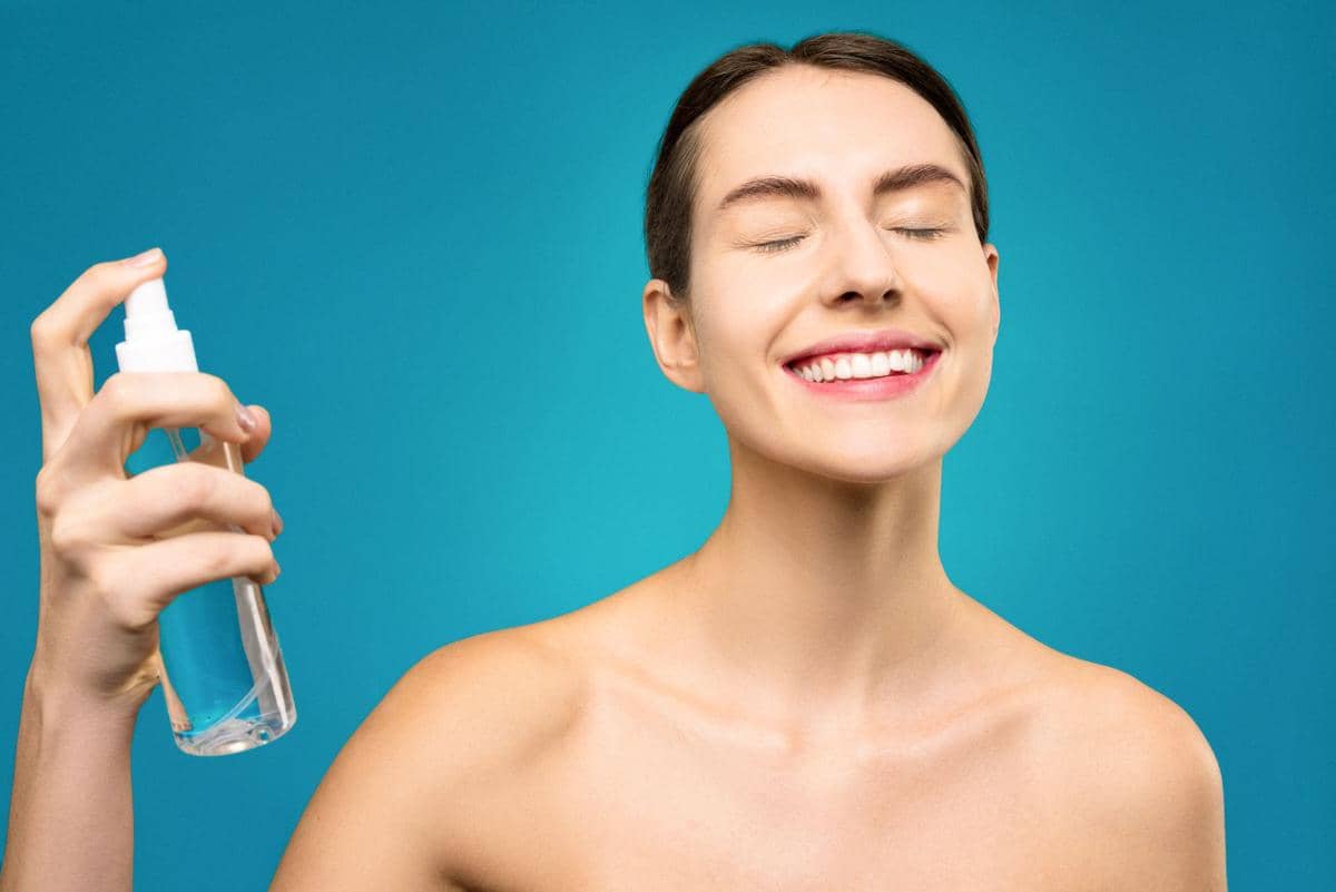 Seorang wanita tersenyum dengan mata tertutup sambil menyemprotkan setting spray ke wajahnya di latar belakang biru.