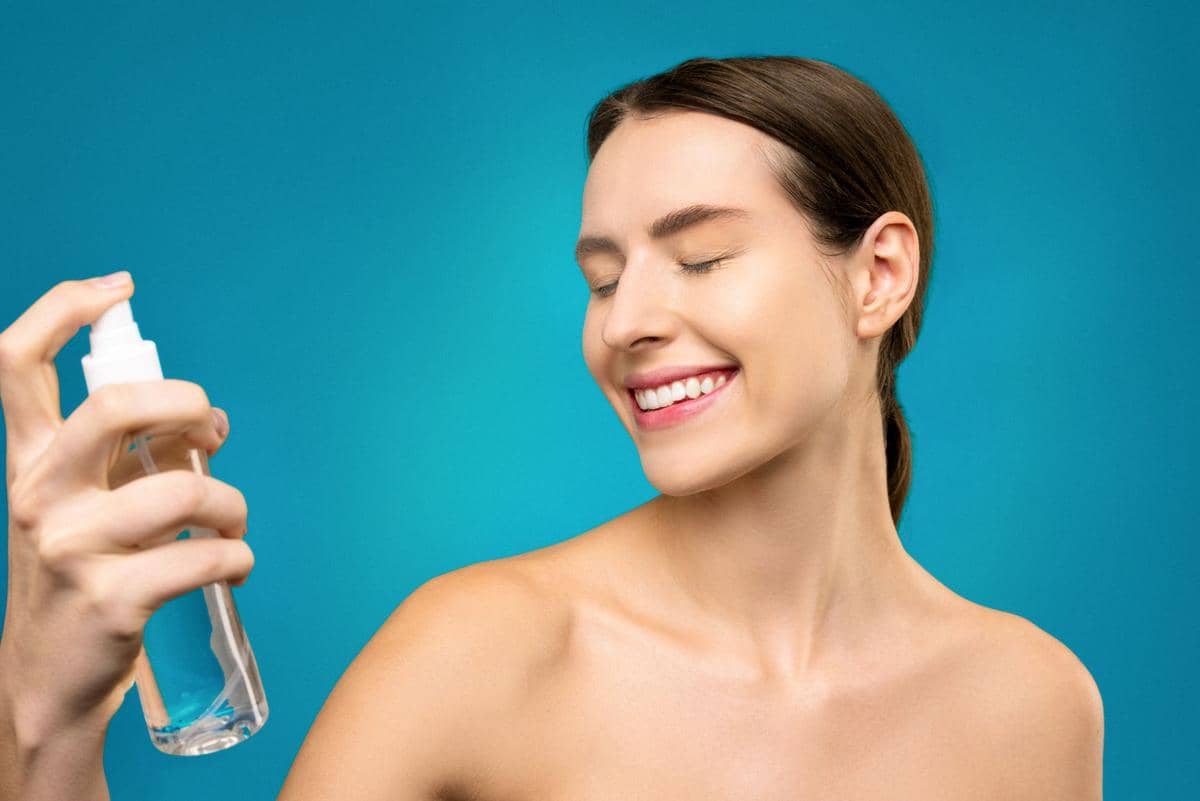 Seorang wanita tersenyum sambil menyemprotkan setting spray ke wajahnya dengan latar belakang biru cerah.