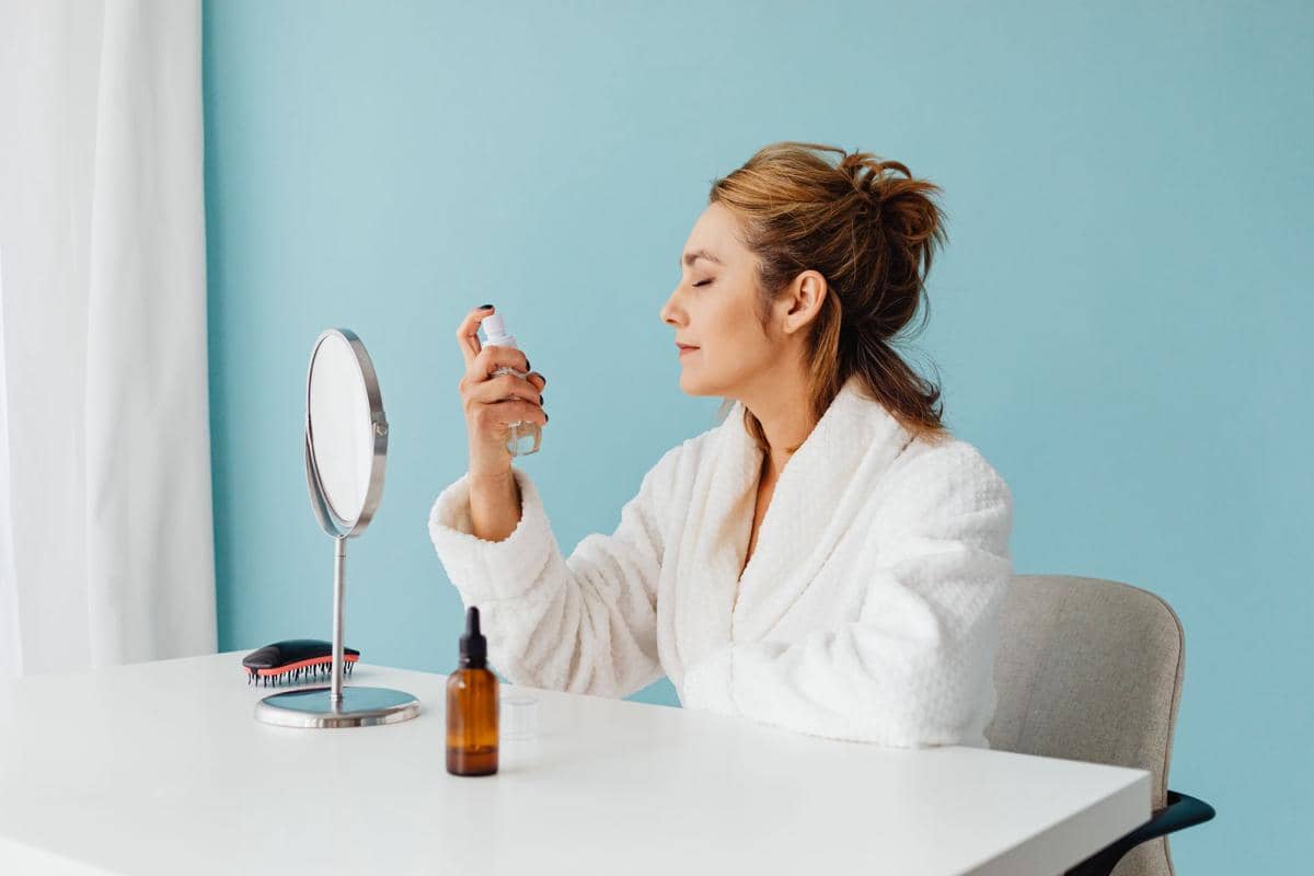 Seorang wanita mengenakan jubah mandi putih menyemprotkan setting spray ke wajahnya di depan cermin di meja rias.