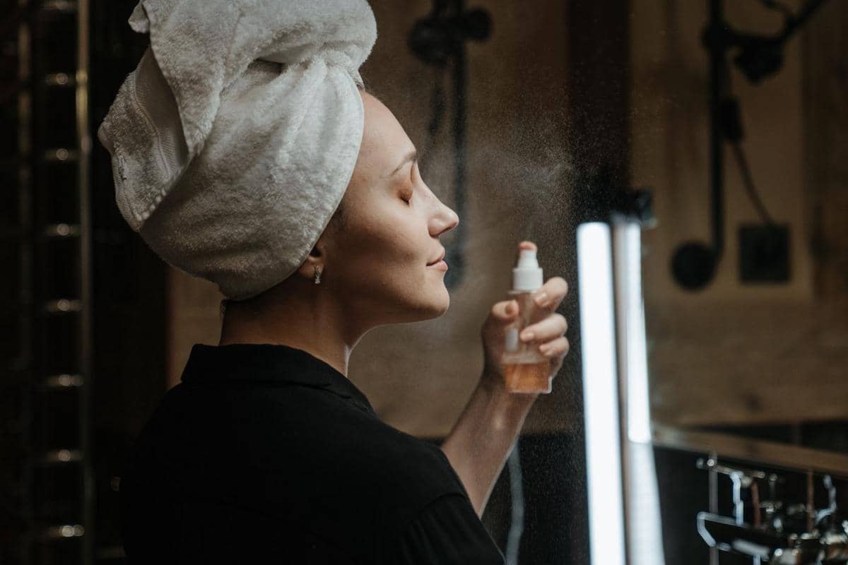 Seorang wanita dengan handuk di kepala menyemprotkan setting spray ke wajahnya di depan cermin dengan pencahayaan lembut.