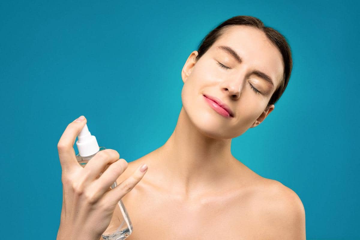 Seorang wanita memejamkan mata sambil menyemprotkan setting spray ke wajahnya dengan latar belakang biru cerah.