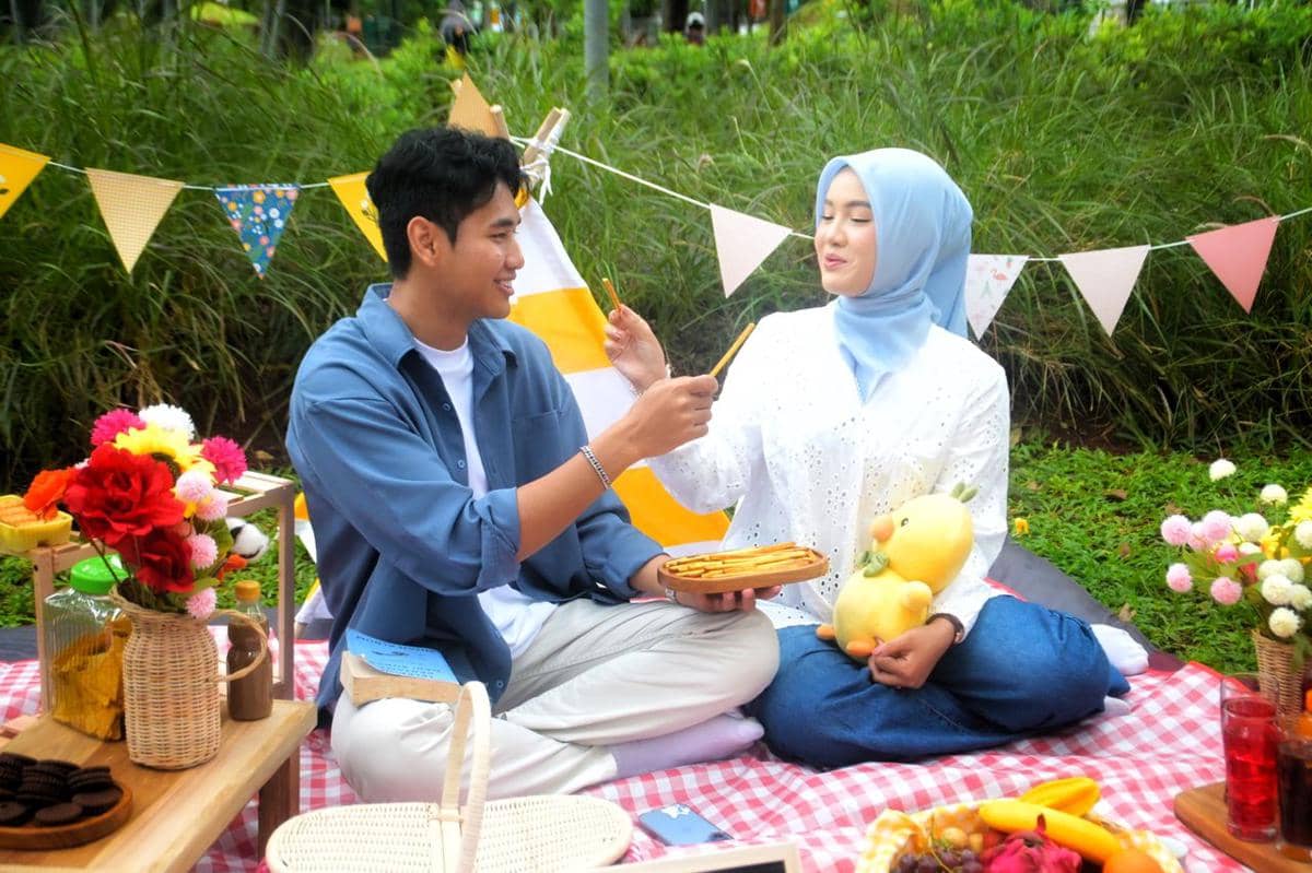 Pasangan muda duduk di atas tikar piknik dengan makanan ringan dan bunga, saling menyuapi camilan di taman hijau.