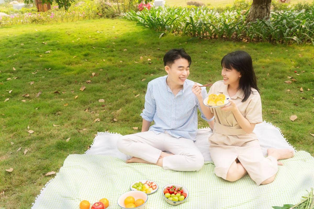 Sepasang muda duduk di atas tikar piknik di taman hijau sambil saling menyuapi makanan dan tersenyum bahagia.