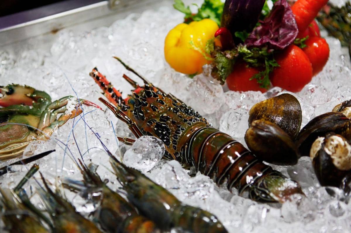 Lobster segar diletakkan di atas es bersama kerang, udang, dan sayuran berwarna cerah seperti paprika dan tomat.