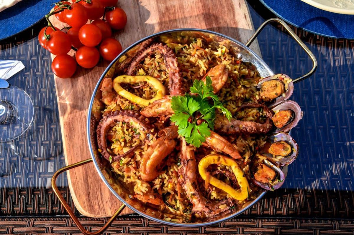 Paella seafood berisi nasi, udang, cumi, gurita, dan kerang disajikan dalam wajan logam dengan hiasan daun peterseli segar.