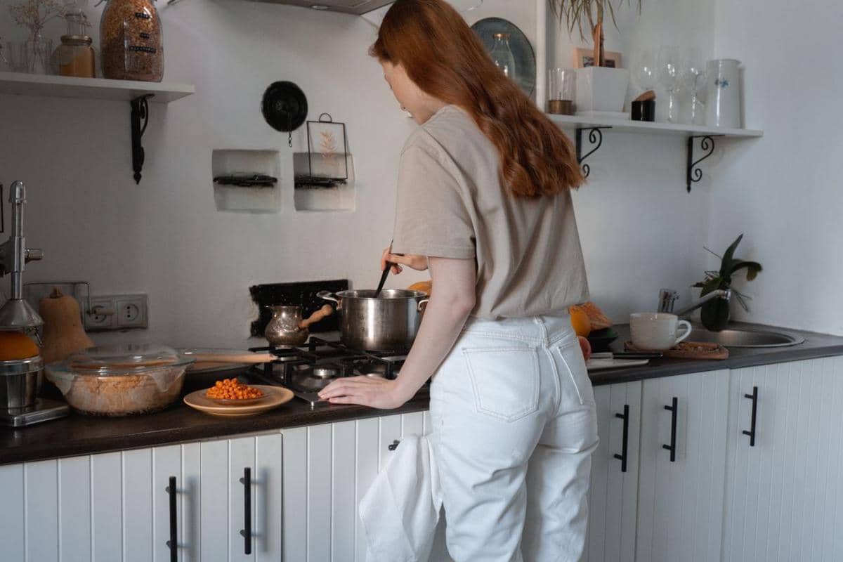 Seorang wanita dengan rambut panjang sedang memasak di dapur modern, mengaduk panci di atas kompor dengan peralatan dapur di sekitarnya.