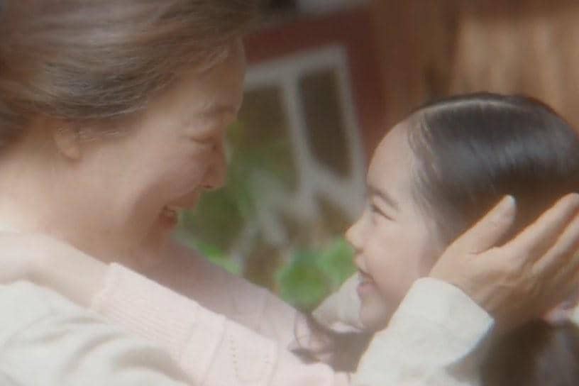 Seorang wanita paruh baya tersenyum hangat sambil memeluk seorang anak perempuan kecil dalam adegan drama My Royal Nemesis.