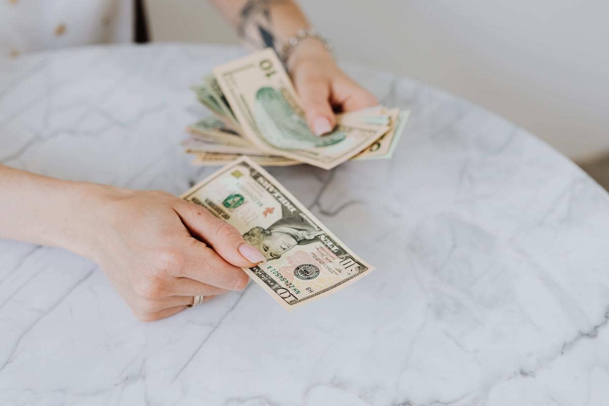 Seseorang memegang dan menghitung lembaran uang dolar Amerika di atas meja marmer berwarna putih.