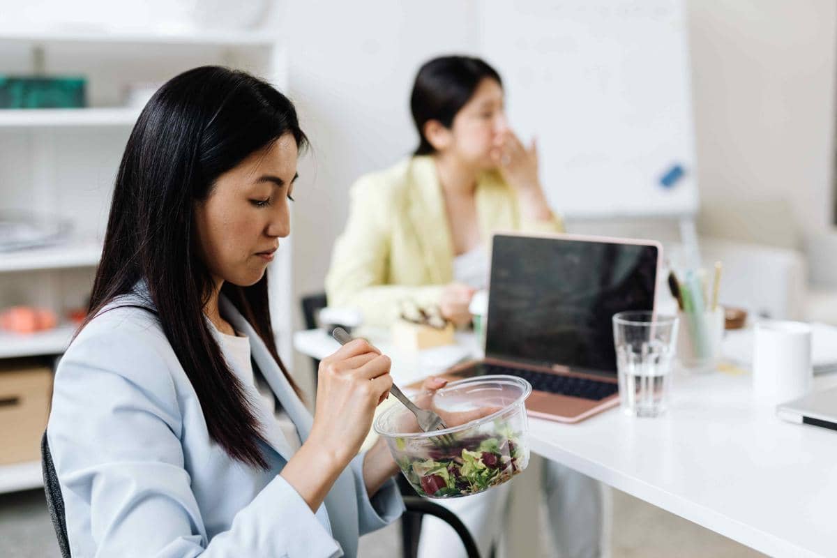 Seorang wanita sedang makan bekal salad di meja kerja dengan laptop terbuka di depannya di lingkungan kantor modern.