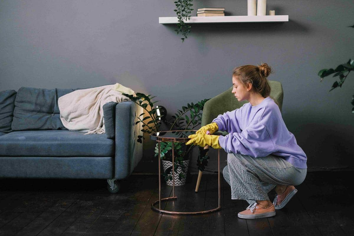 Seorang wanita mengenakan sarung tangan karet kuning sedang membersihkan meja kecil di ruang tamu dengan sofa biru dan tanaman hias.