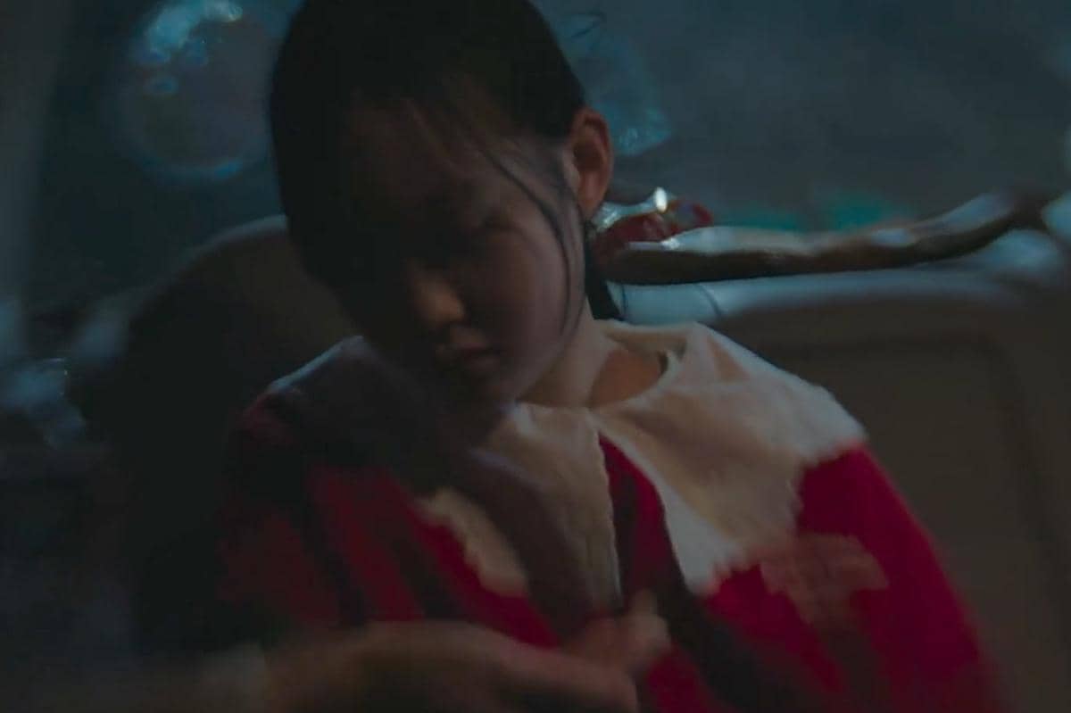 Seorang anak perempuan mengenakan kostum merah dengan kerah putih duduk di dalam mobil dalam suasana gelap dan suram.