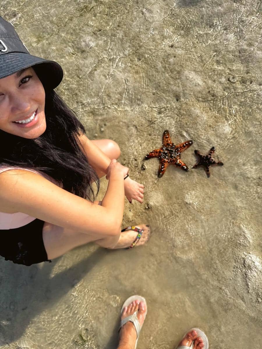 Seorang perempuan tersenyum duduk di tepi air dangkal bersama dua bintang laut berwarna oranye di Telaga Nirwana, Nusa Tenggara Timur.