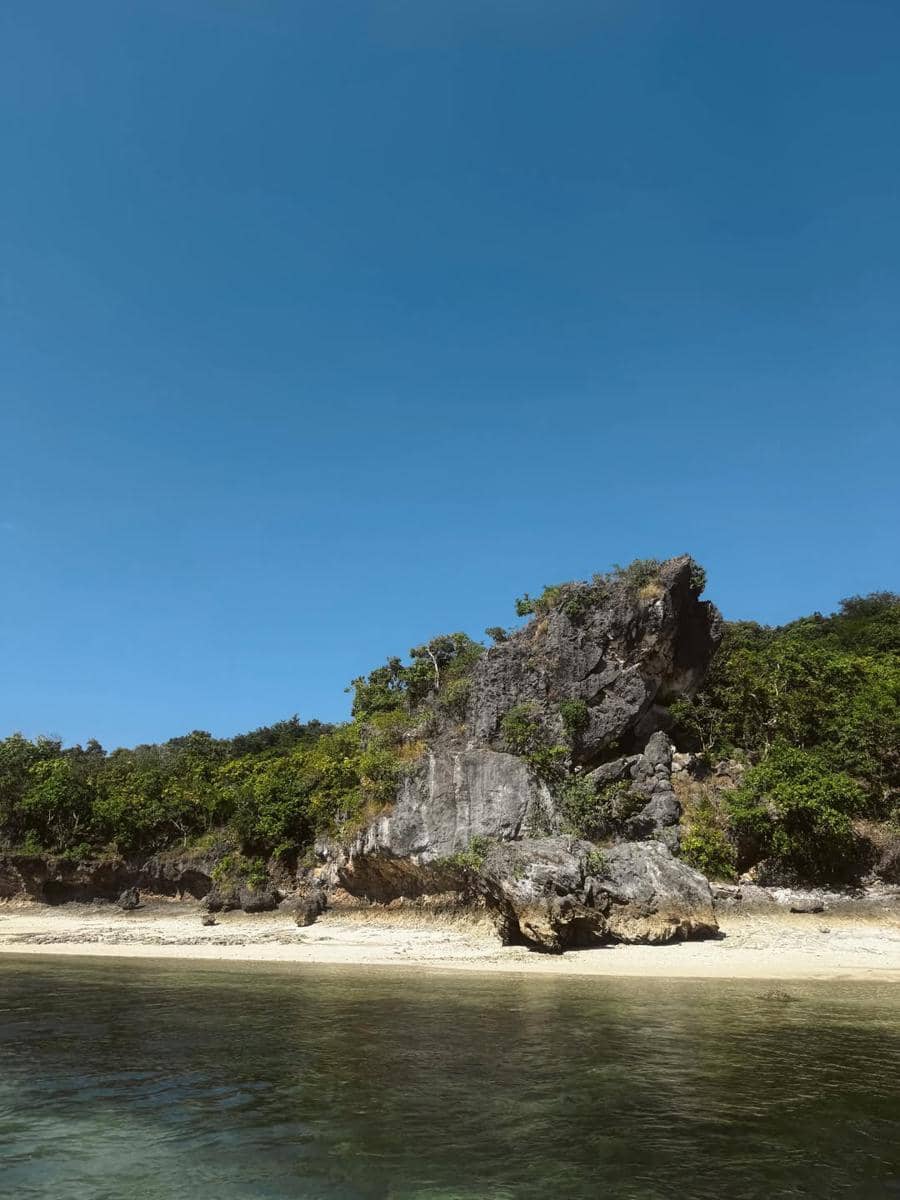 Pemandangan tebing batu dan pantai berpasir putih di Telaga Nirwana, Nusa Tenggara Timur, dengan air laut jernih dan langit biru cerah.