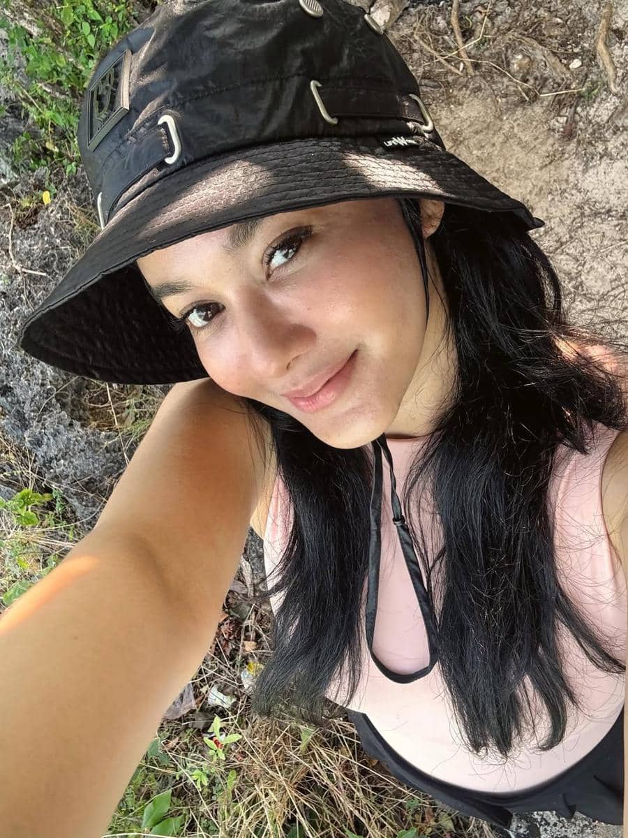 Seorang perempuan berambut hitam mengenakan topi hitam dan pakaian tanpa lengan berwarna muda berpose selfie di area alam terbuka.