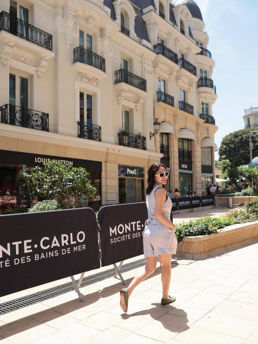 Seorang wanita mengenakan pakaian musim panas berjalan di area perbelanjaan Monte Carlo, Monaco dengan latar bangunan bergaya Eropa klasik.