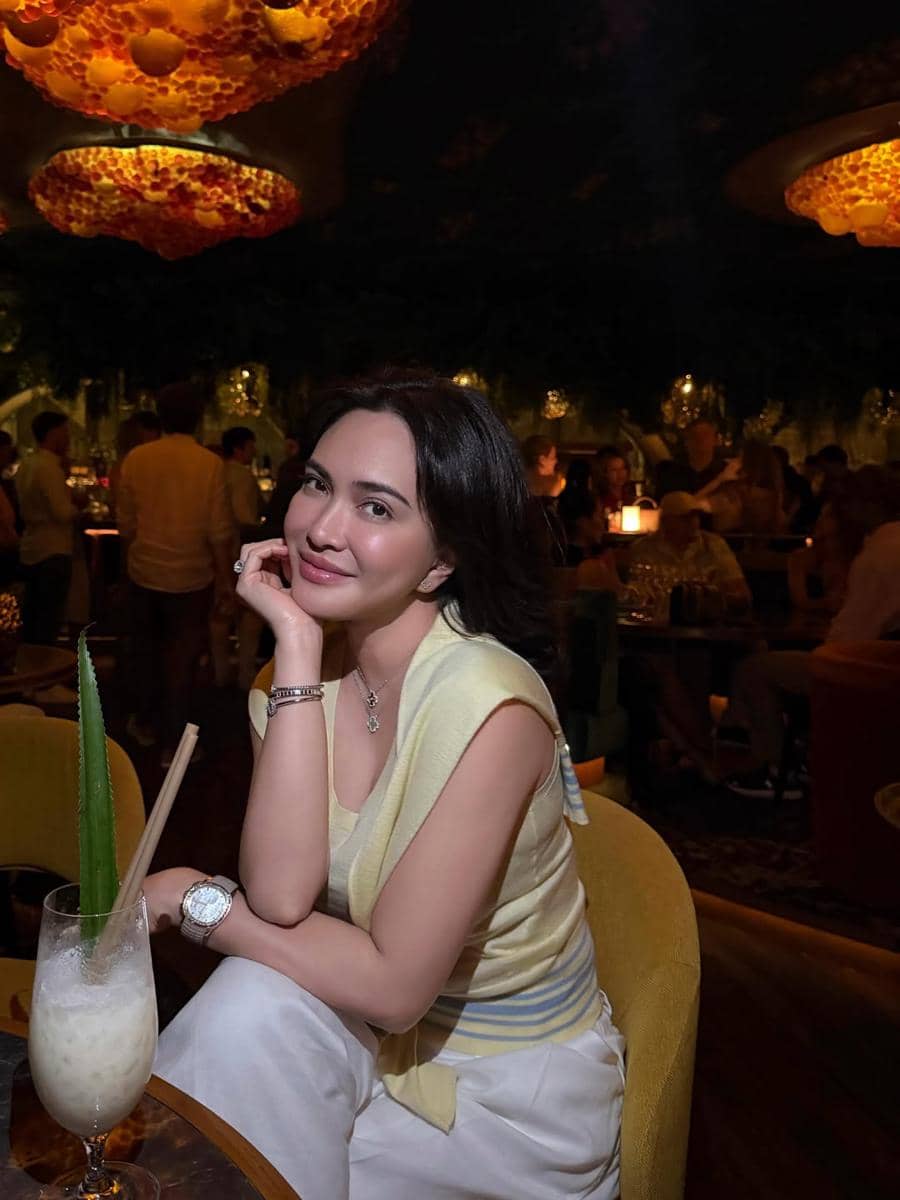 Seorang wanita berpose santai di dalam restoran mewah dengan pencahayaan hangat, mengenakan pakaian kasual berwarna lembut dan minuman di mejanya.