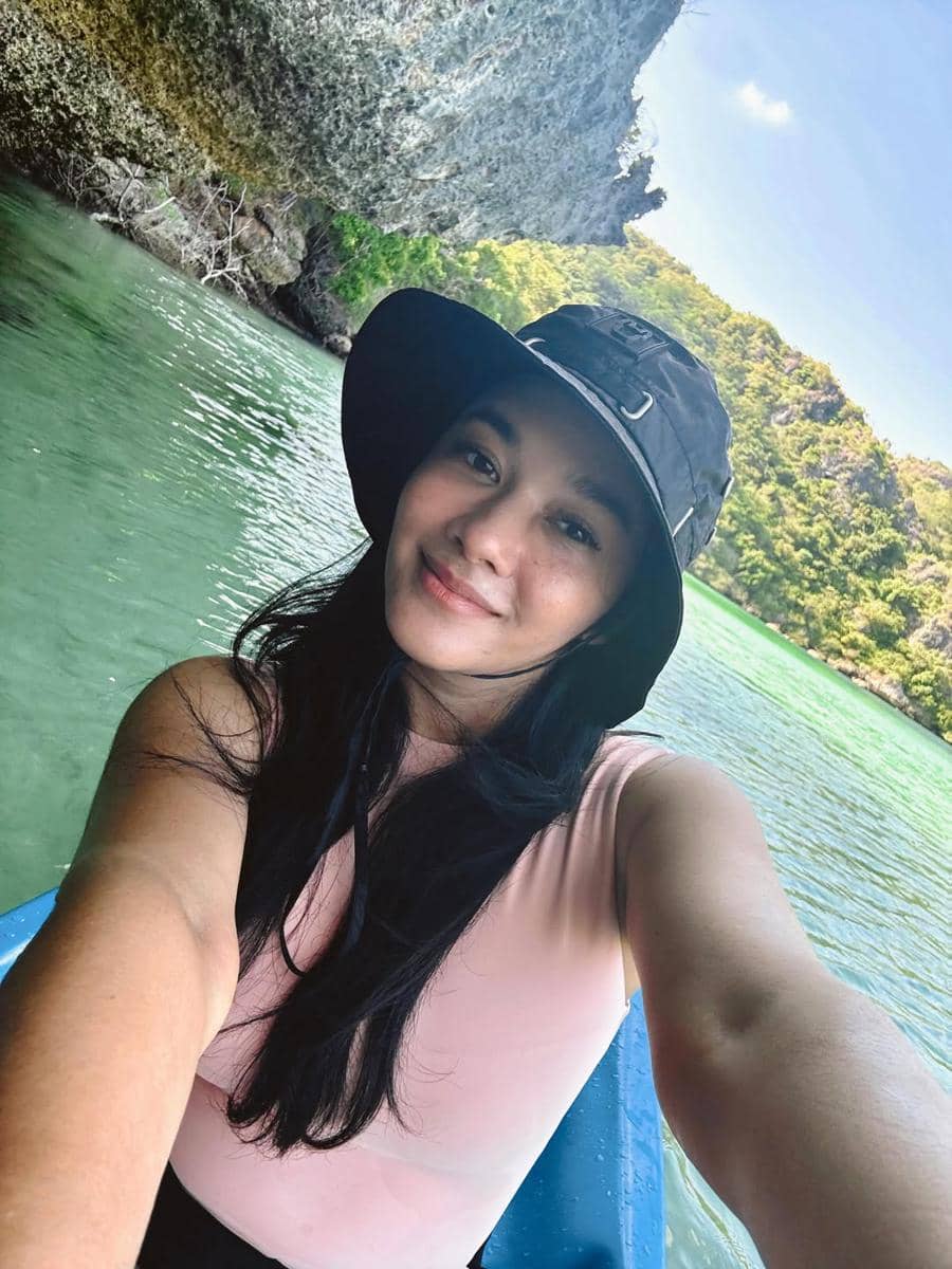 Seorang perempuan berpose selfie di atas perahu di Telaga Nirwana, Nusa Tenggara Timur, dengan air hijau jernih dan tebing batu di sekitarnya.