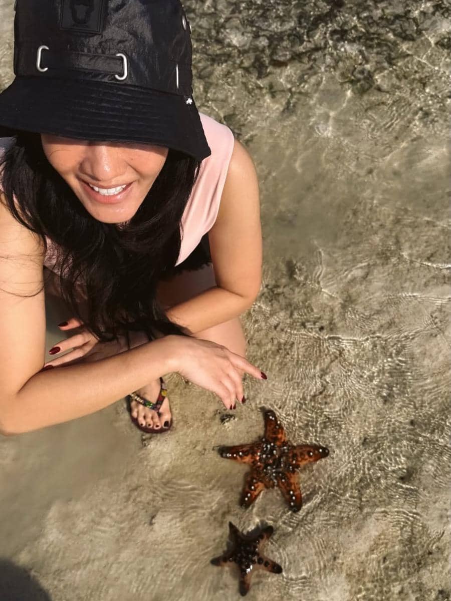 Seorang perempuan tersenyum sambil berjongkok di tepi air jernih, menunjuk dua bintang laut berwarna cokelat di Telaga Nirwana, NTT.