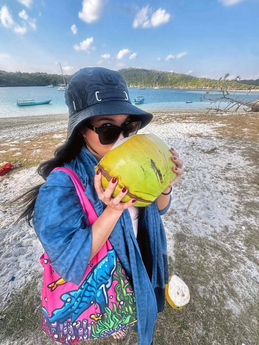 Seorang perempuan mengenakan topi dan kacamata hitam menikmati air kelapa di tepi Telaga Nirwana, Nusa Tenggara Timur, dengan latar perahu dan perbukitan hijau.