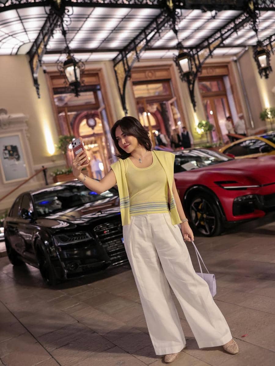 Seorang wanita berpose sambil berswafoto di depan gedung mewah dengan mobil sport hitam dan merah terparkir di Monaco pada malam hari.