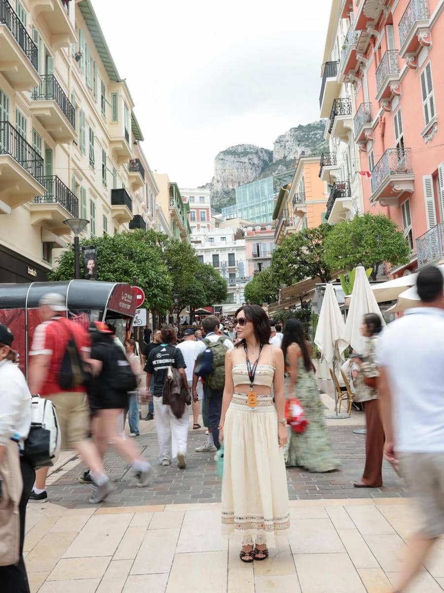 Seorang wanita mengenakan gaun krem berdiri di jalan ramai dengan latar bangunan warna-warni dan pegunungan di Monaco.