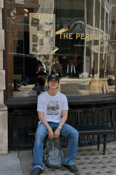 Seorang pria mengenakan kaus putih, celana jeans, dan topi hitam duduk di bangku depan toko bertuliskan THE PERFUMER di area perkotaan.