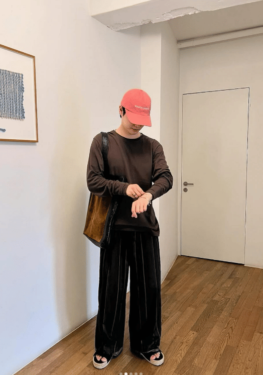 Seorang pria mengenakan topi merah muda, kaus lengan panjang cokelat tua, celana panjang hitam longgar, dan sandal di ruangan minimalis.