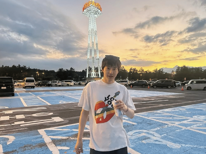 Seorang pria mengenakan kaus putih bergambar hati dan memegang minuman berdiri di area parkir dengan latar menara bercahaya saat matahari terbenam.