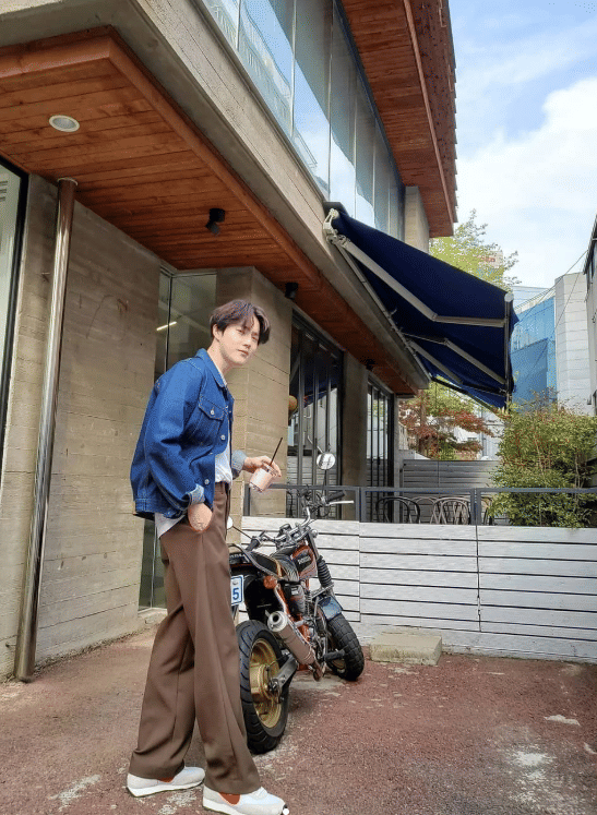 Seorang pria berpose santai di depan bangunan modern dengan motor kecil di sampingnya, mengenakan jaket denim dan celana cokelat.