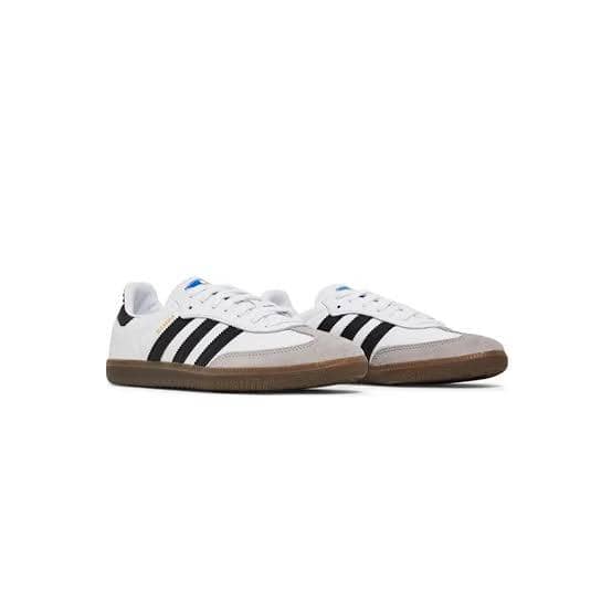 Sepasang sepatu Adidas Samba berwarna putih dengan tiga garis hitam dan sol karet cokelat ditampilkan di atas latar putih.