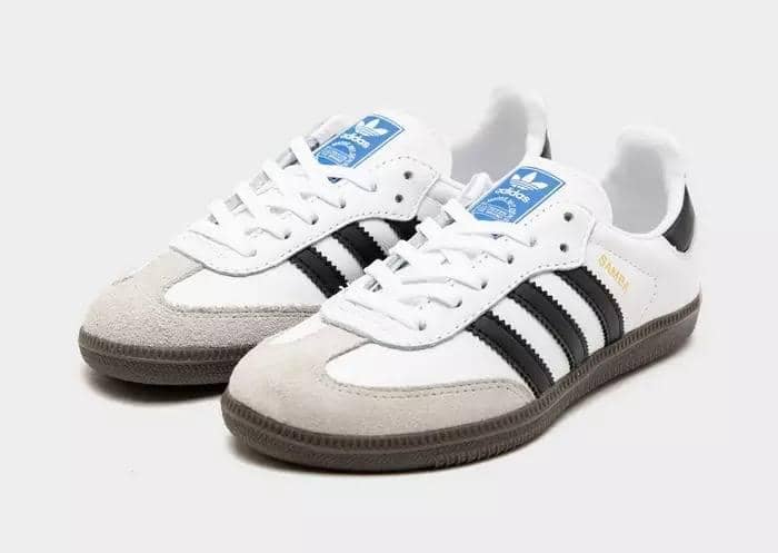 Sepasang sepatu Adidas Samba berwarna putih dengan aksen hitam dan abu-abu, menampilkan desain klasik dengan sol karet cokelat.