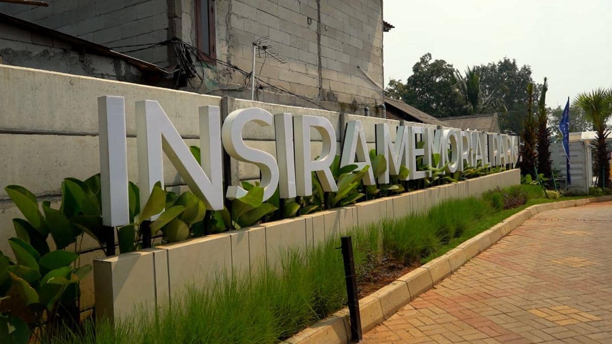 Tampak area depan Insira Memorial Park dengan tulisan besar berwarna putih di dinding taman dan jalan berpaving di sekitarnya.