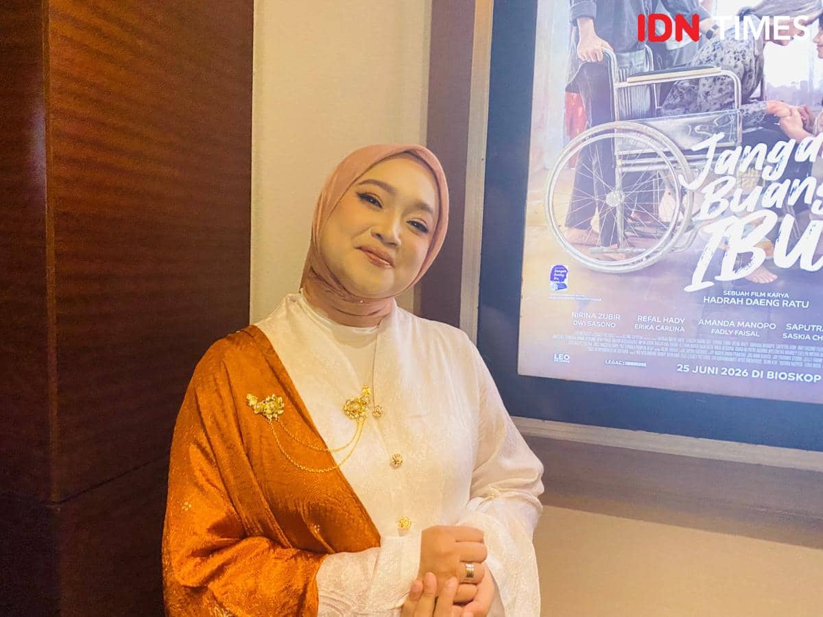 Seorang perempuan berhijab berpose di depan poster film berjudul Jangan Buang Ibu yang menampilkan kursi roda dan dua tokoh lanjut usia.