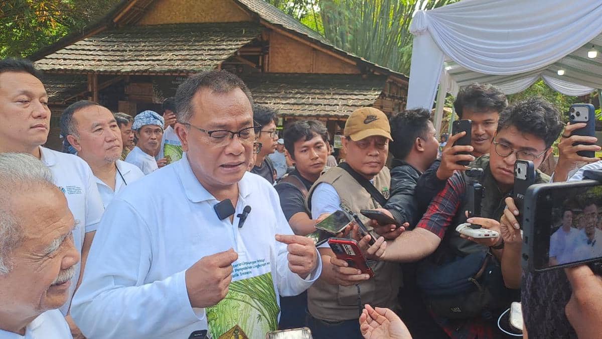 Seorang pria berbaju putih berbicara kepada wartawan yang mengerumuninya sambil memegang ponsel dan alat perekam di area luar ruangan.