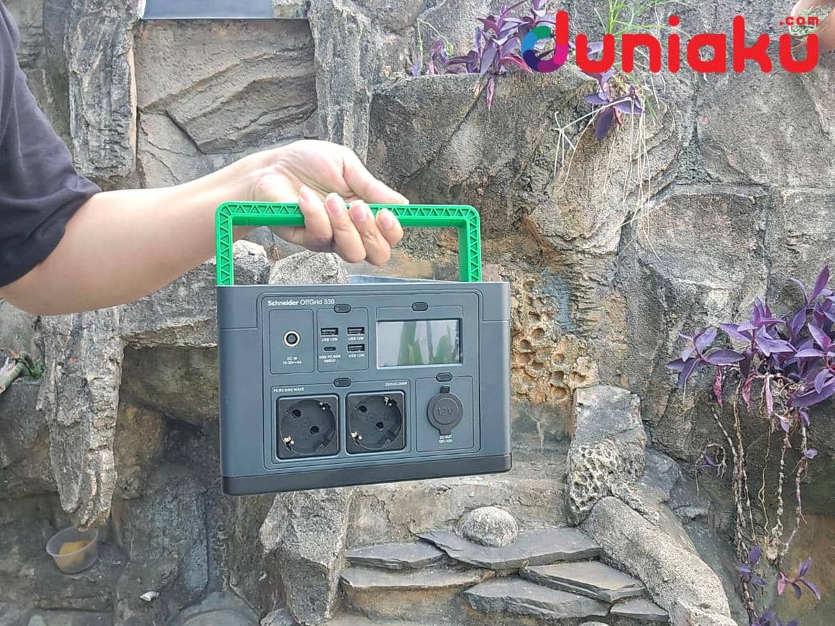 Seseorang memegang Schneider OffGrid Portable Power Station dengan pegangan hijau di area luar ruangan berbatu.