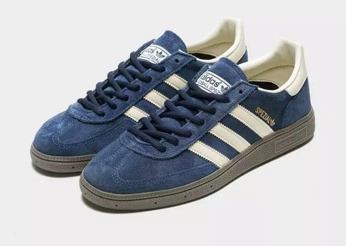 Sepasang sepatu Adidas Spezial berwarna biru dengan tiga garis putih di sisi dan sol abu-abu, diletakkan di permukaan terang.