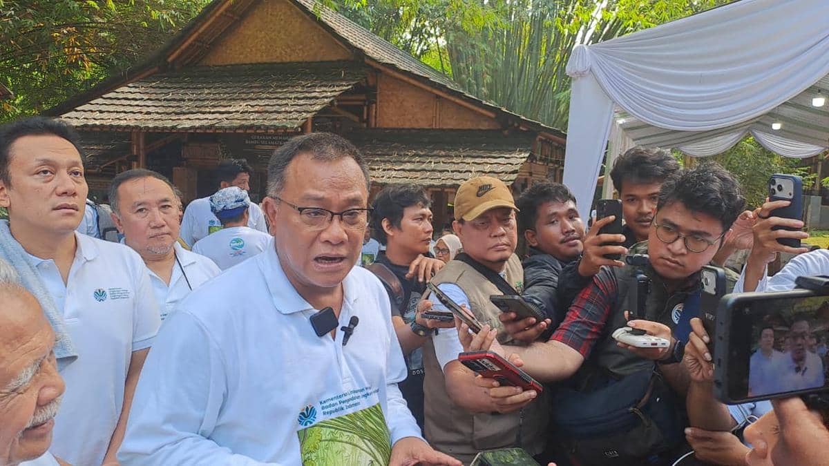 Seorang pejabat mengenakan kemeja putih berbicara kepada wartawan yang mengerumuninya sambil memegang ponsel dan alat perekam di area terbuka.