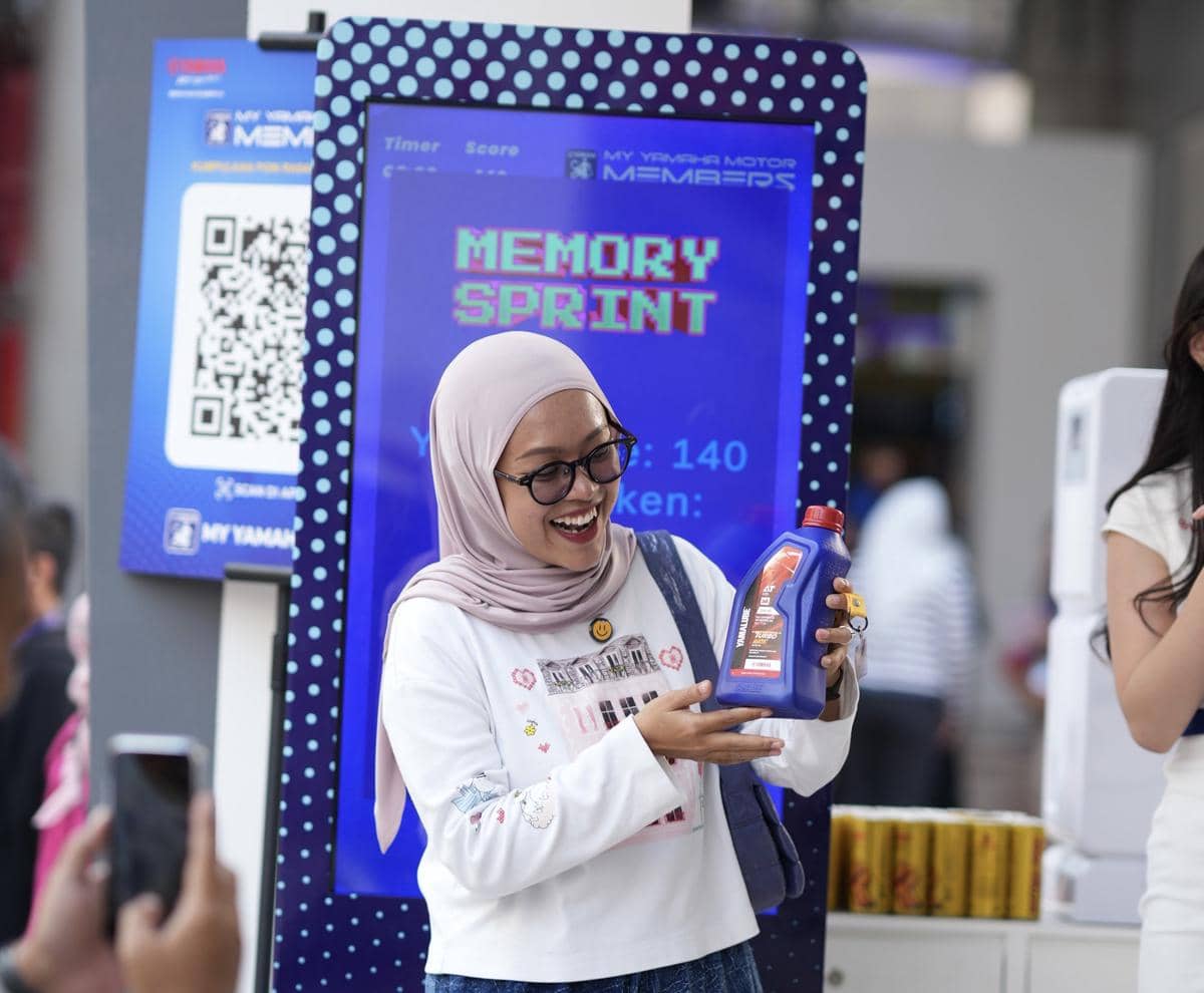 Seorang wanita berhijab tersenyum sambil memegang botol oli di depan layar permainan interaktif dengan latar suasana acara promosi.