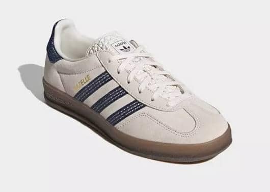 Sepatu Adidas Gazelle berwarna krem dengan tiga garis biru tua di sisi dan sol karet cokelat bergaya klasik.