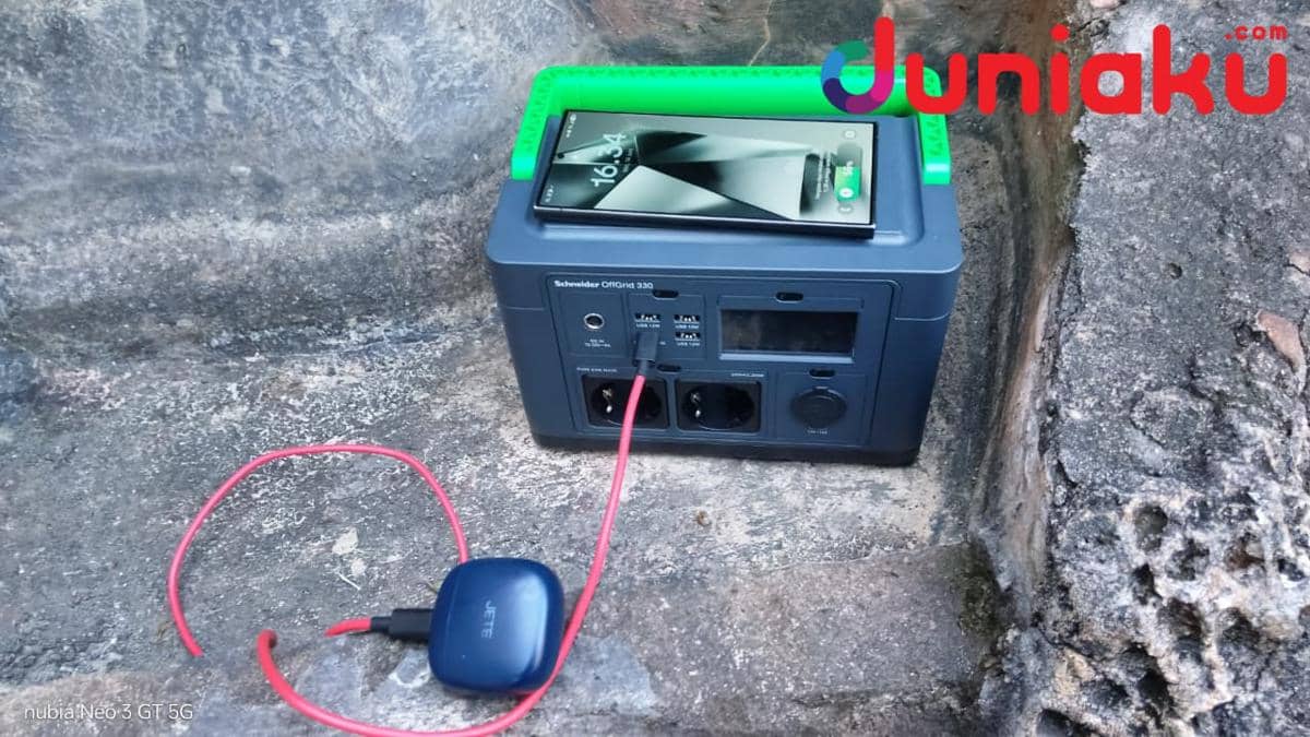 Perangkat Schneider OffGrid sedang mengisi daya smartphone dan earphone TWS dengan kabel merah di area berbatu.