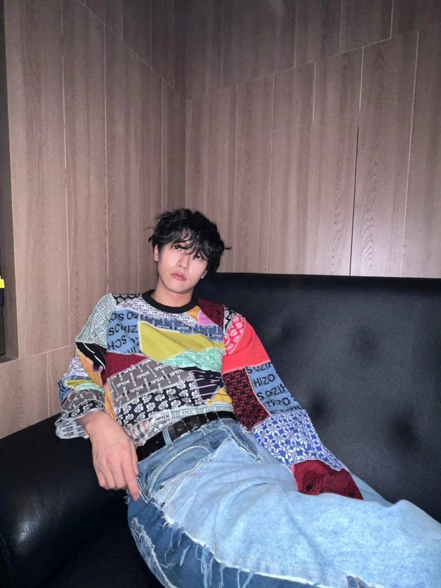 Seorang pria berambut hitam duduk santai di sofa hitam, mengenakan sweater warna-warni bermotif patchwork dan celana jeans robek.