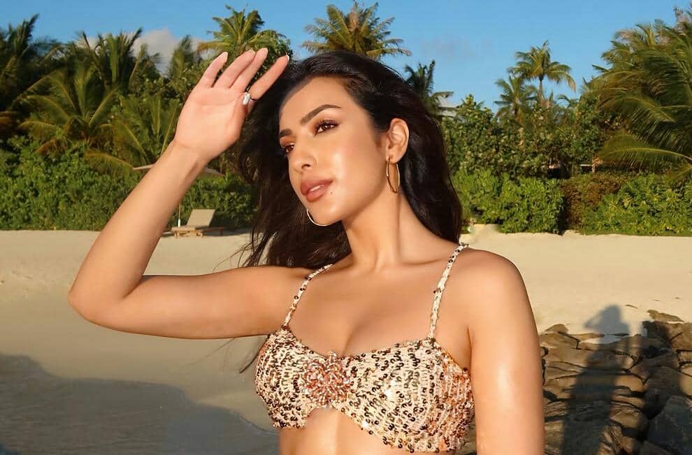 Seorang wanita mengenakan bikini bermotif leopard berpose di pantai berpasir putih dengan latar belakang pepohonan tropis dan langit cerah.